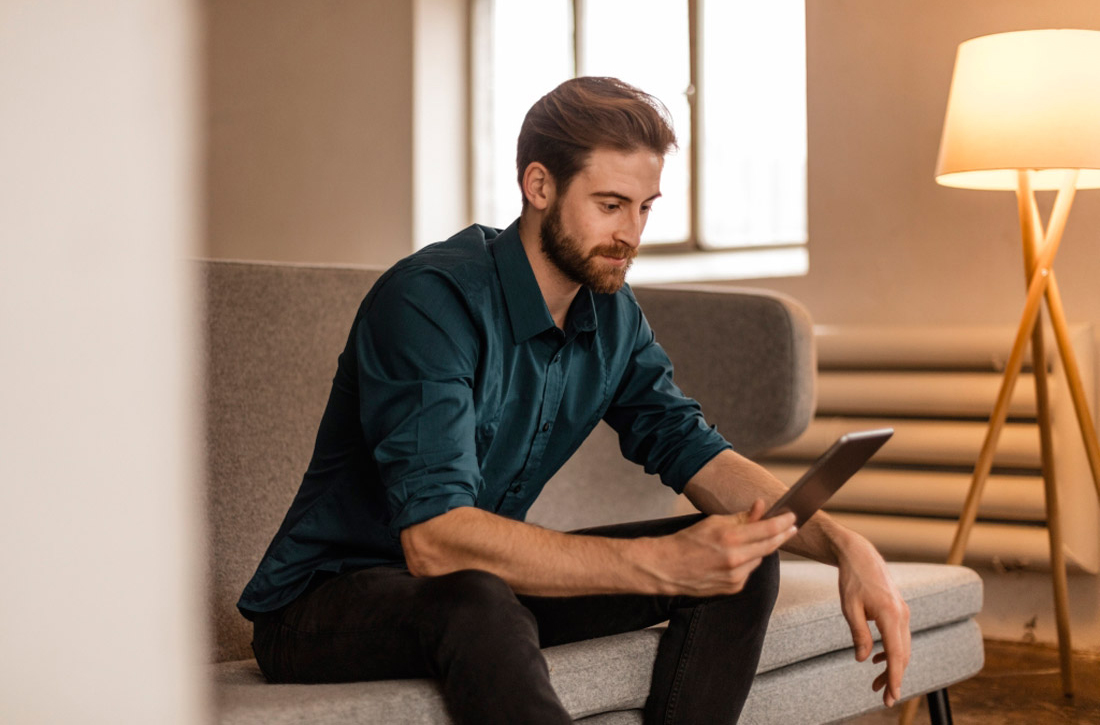 Ein Mann sitzt im hell erleuchteten Wohnzimmer auf dem Sofa und schaut auf sein Tablet.