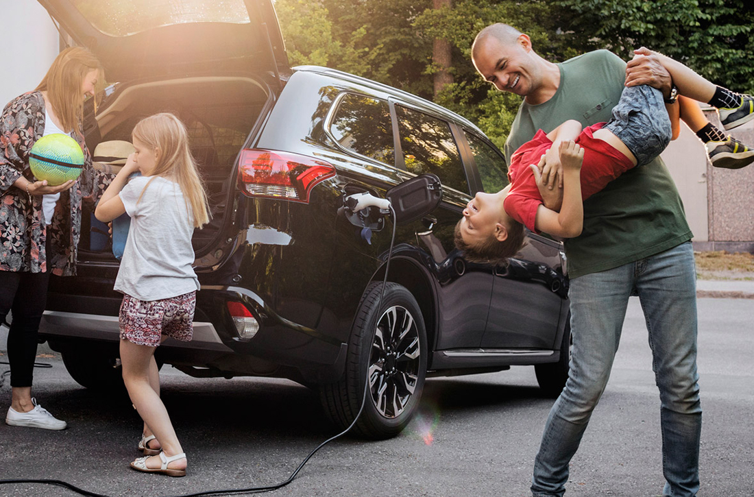 Familie steht zuhause an einem Elektroauto, Vater und Sohn spielen.