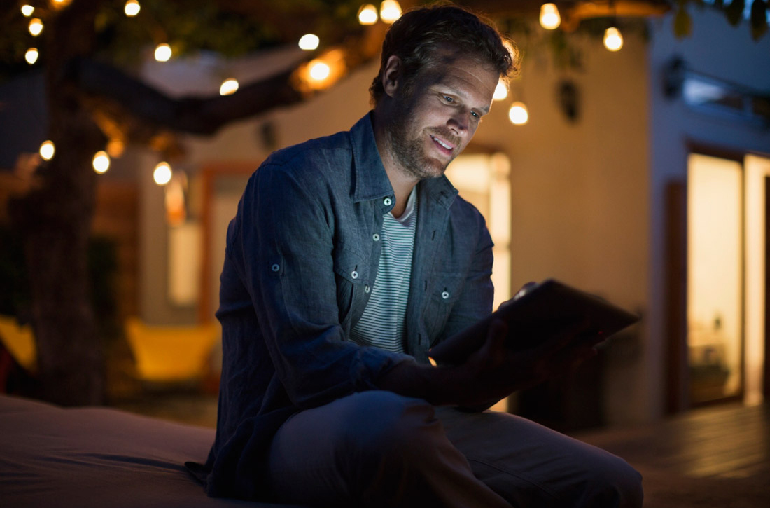 Ein Mann sitzt bei Dunkelheit draußen in einem mit Lichterketten erleuchteten Garten und schaut auf sein Tablet.