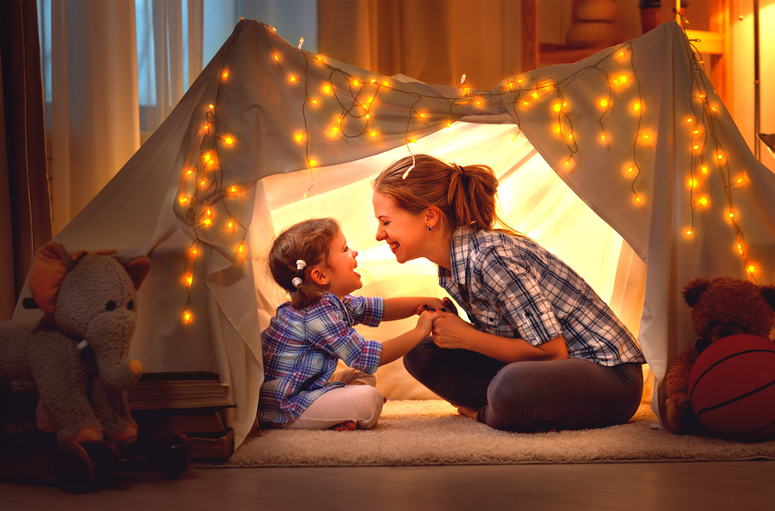 Mutter sitzt mit ihrer kleinen Tochter in einem mit einer Lichterkette erleuchteten Zelt im Kinderzimmer. 